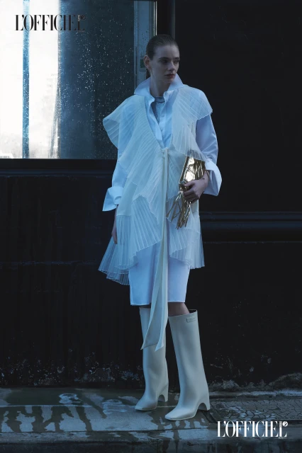 Fashion editorial shot of a fair-skinned adult female model wearing a ruffled top, shorts, and boots, holding a gold clutch.