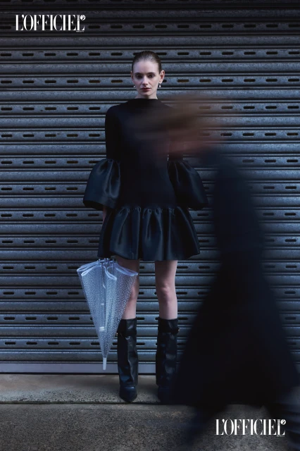 A slender woman poses in a black dress with an umbrella against a gray roller door. Her dress has bell sleeves and a ruffled skirt.