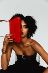 Black woman holding a glossy red handbag up to her face against a white seamless studio background