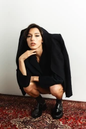 Woman in a black blazer draped over her shoulders, crouching on an oriental rug against a plain white wall