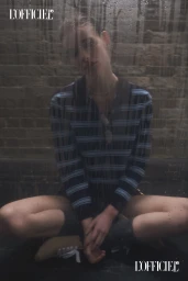 A woman in a navy striped polo sweater sits cross-legged against a brick wall, viewed through rain-streaked glass.