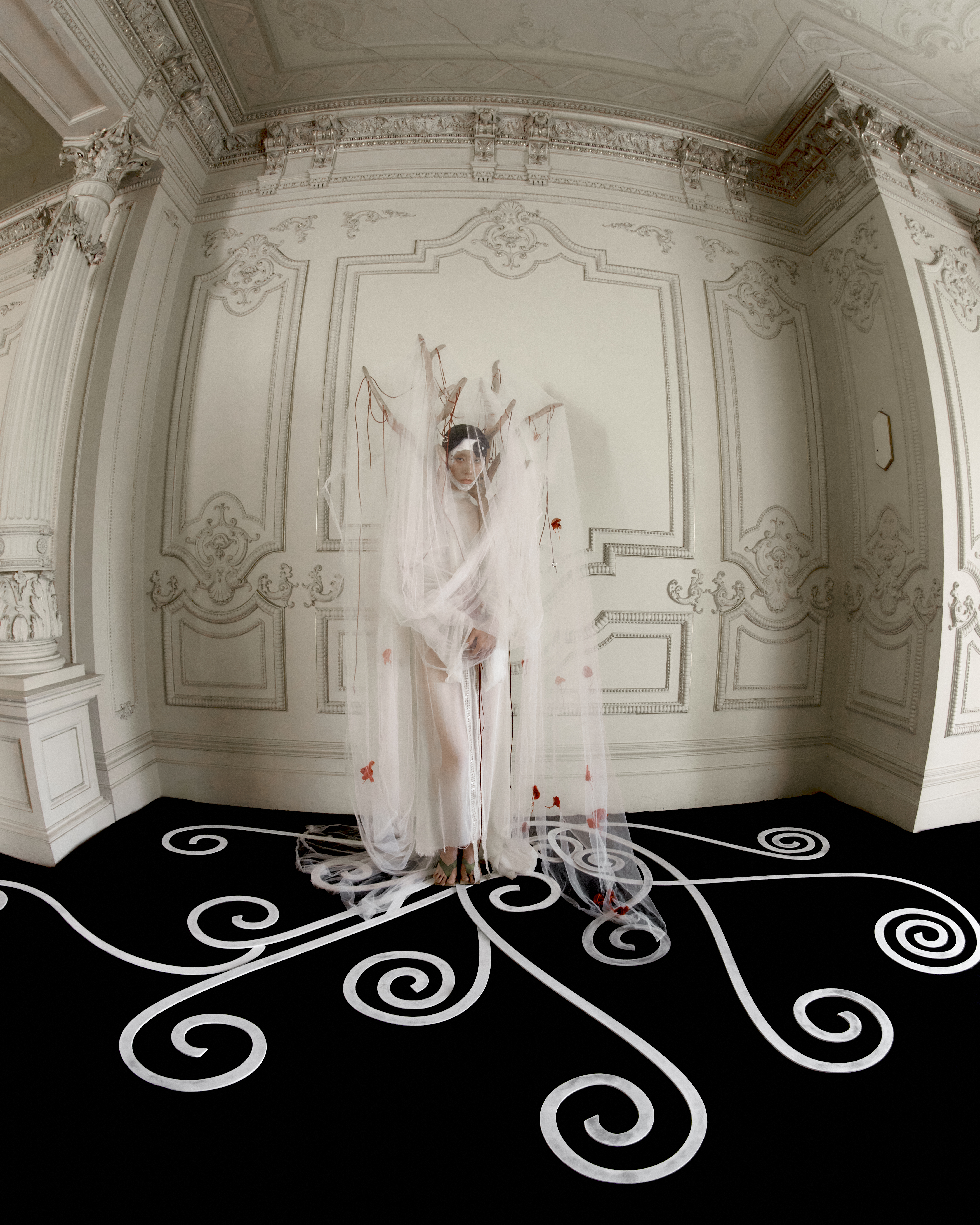 Indoor portrait of a person in a white veil with red flower accents, standing on a black floor with white decorative swirls.