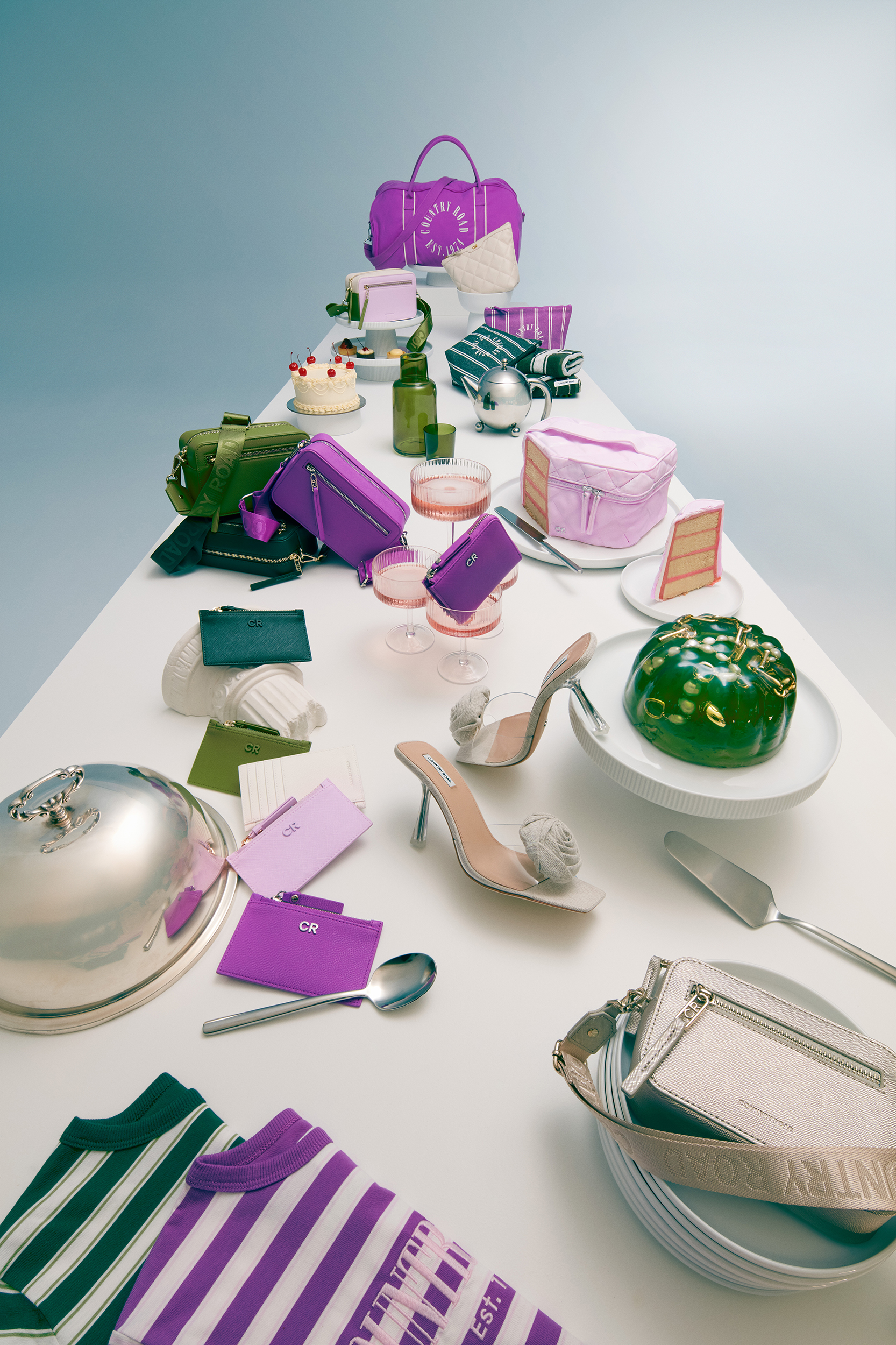 Arrangement of colorful handbags, purses, shoes, cakes, and tableware on a white table against a light blue background.