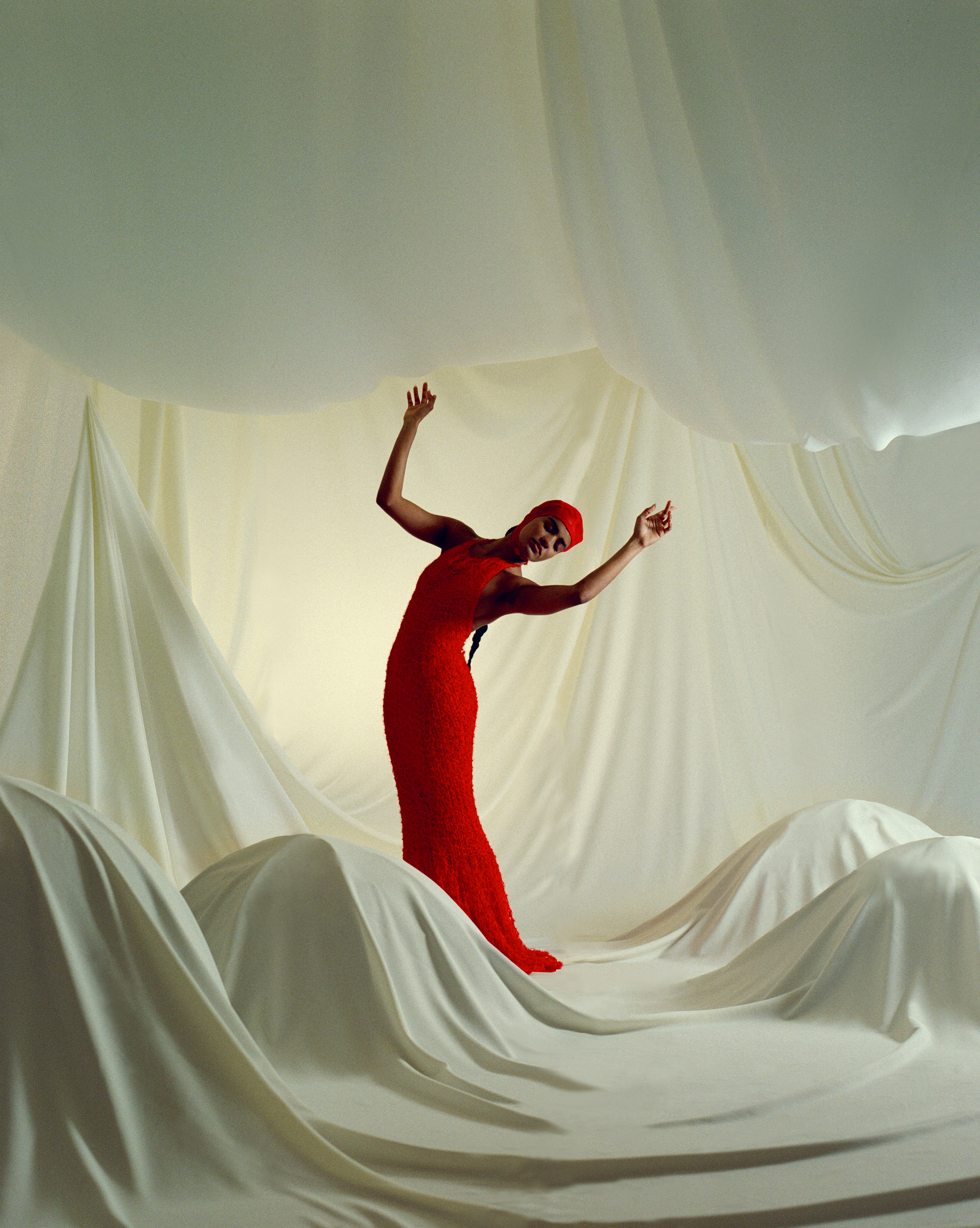 An adult female in a red dress bends backward on a white draped surface with white draped fabrics overhead in a studio setting.