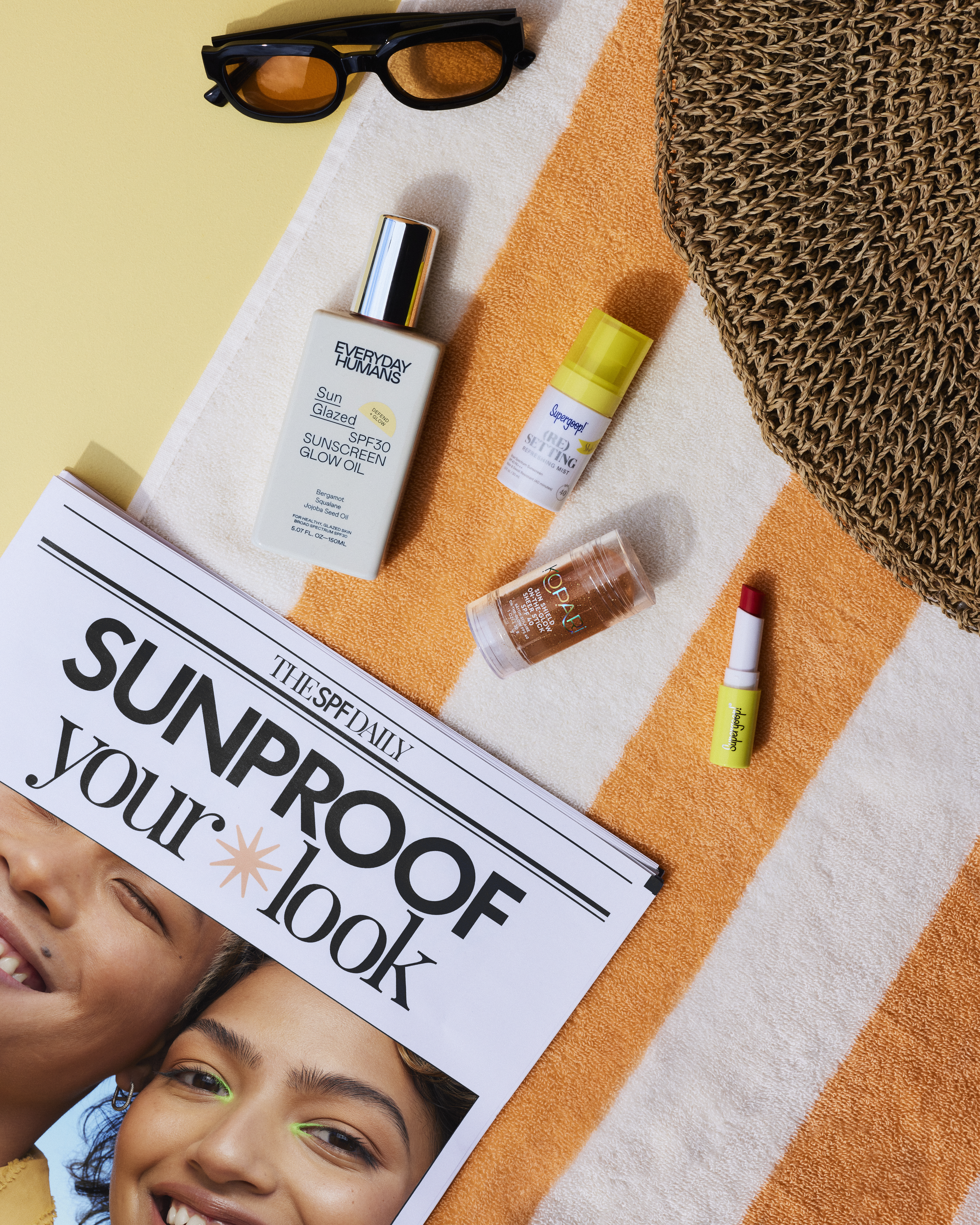 Flat lay of skincare products, sunglasses, newspaper, and beach bag on an orange and white striped towel against a yellow backdrop.