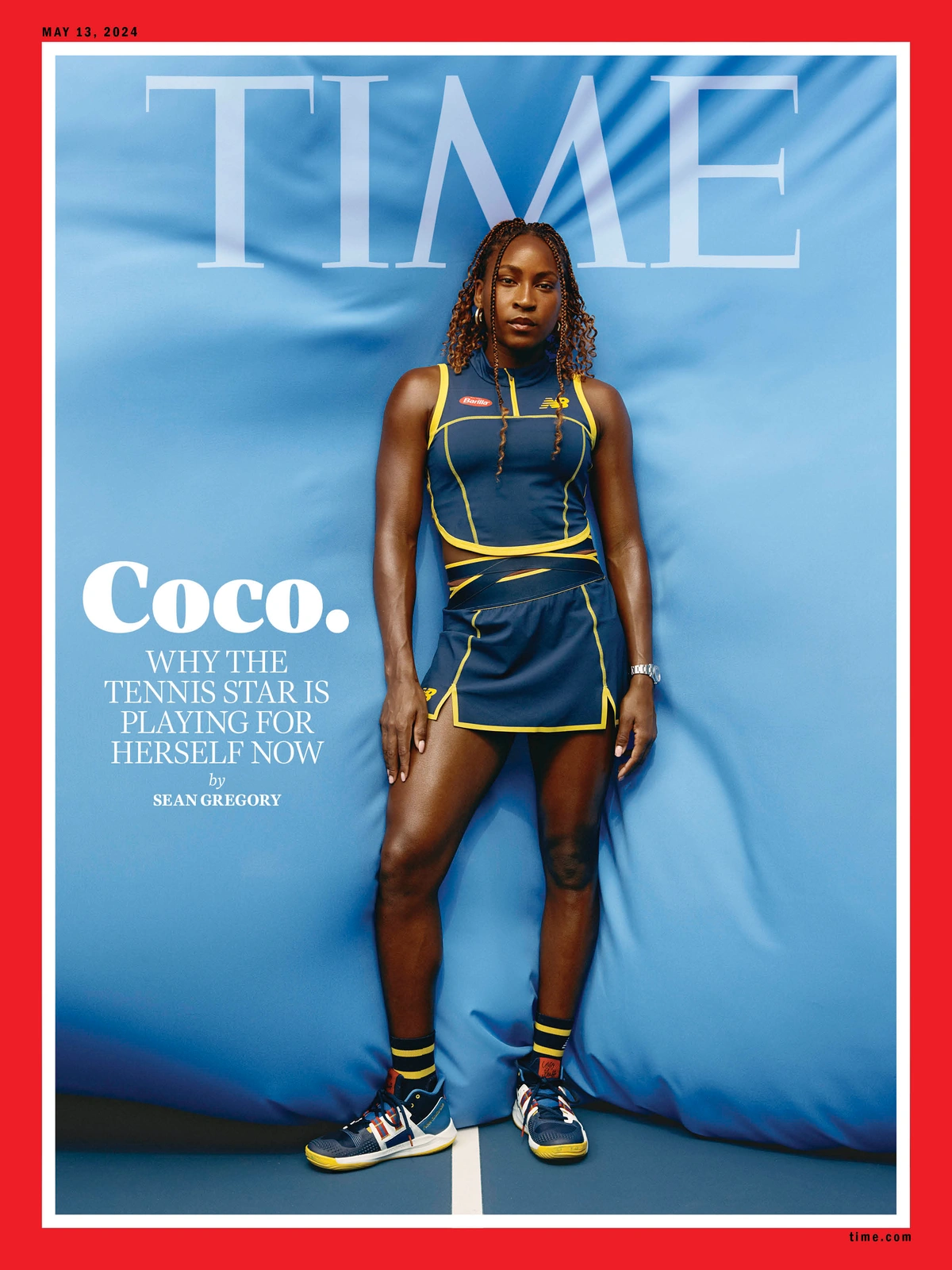 Coco Gauff standing against a bright blue fabric backdrop wearing a navy athletic top and matching skirt with yellow trim