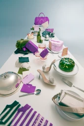 A long white table displaying assorted purple bags, striped sweaters, cakes, and high-heel shoes.