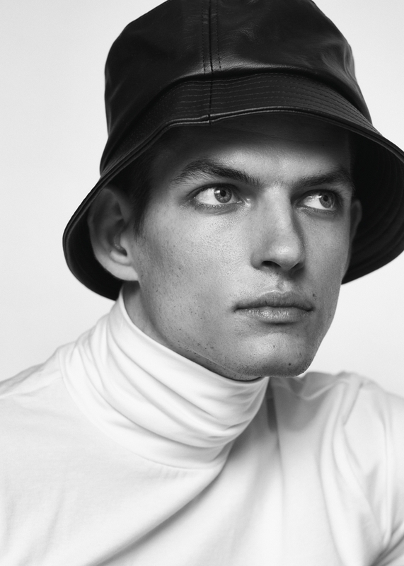 Monochrome portrait of an adult male wearing a black leather bucket hat and white turtleneck sweater against a solid light background.