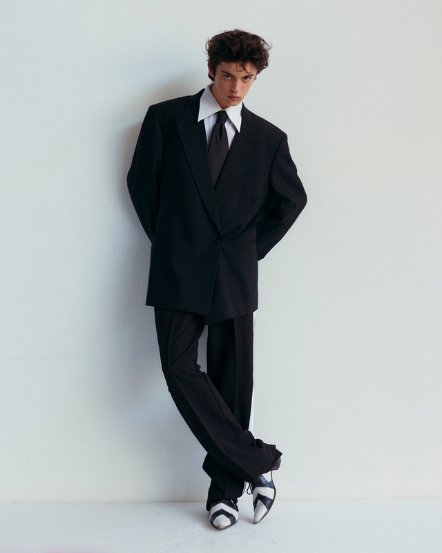 A young adult male fashion model in a black double-breasted suit and tie, posing in front of a white wall in a studio.