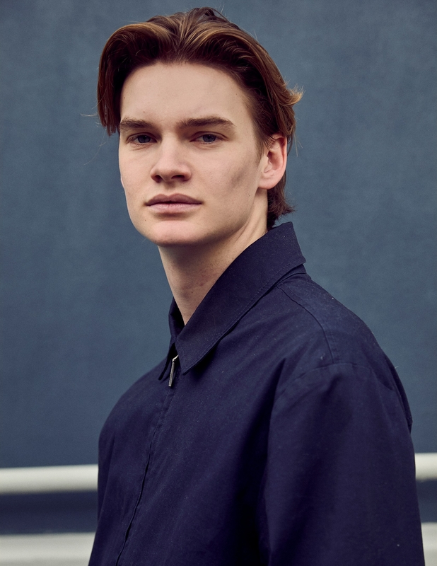 Portrait of a young adult male with light skin and brown hair wearing a navy blue zip-up jacket against a solid blue background.