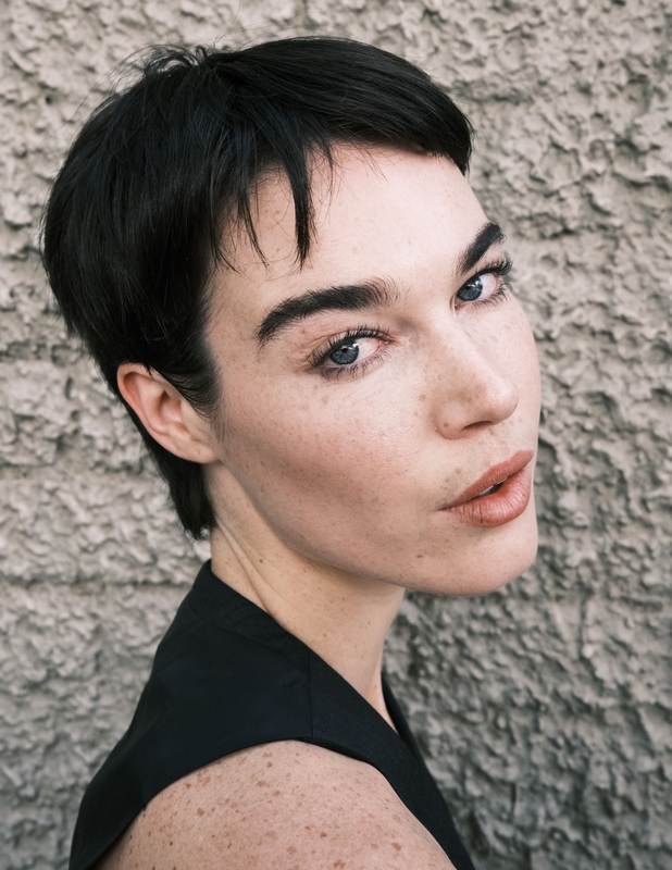 Close-up portrait of a fair-skinned adult female with a dark pixie cut and black top, posing against a textured grey wall backdrop.