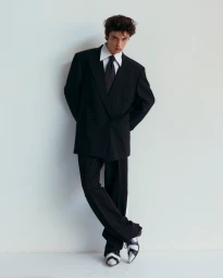 Young man in a black oversized suit jacket, white shirt, black tie and two tone shoes leaning against a plain white wall