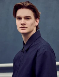 Close-up portrait of a young male model with brown hair wearing a dark blue collared jacket against a textured blue wall.