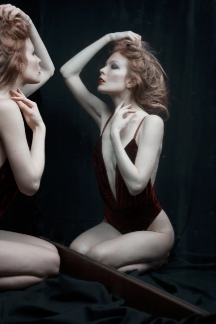 An adult woman poses in a burgundy swimsuit near a mirror, her hand in her hair, against a dark background in a studio setting.