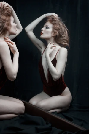 An adult woman poses in a burgundy swimsuit near a mirror, her hand in her hair, against a dark background in a studio setting.