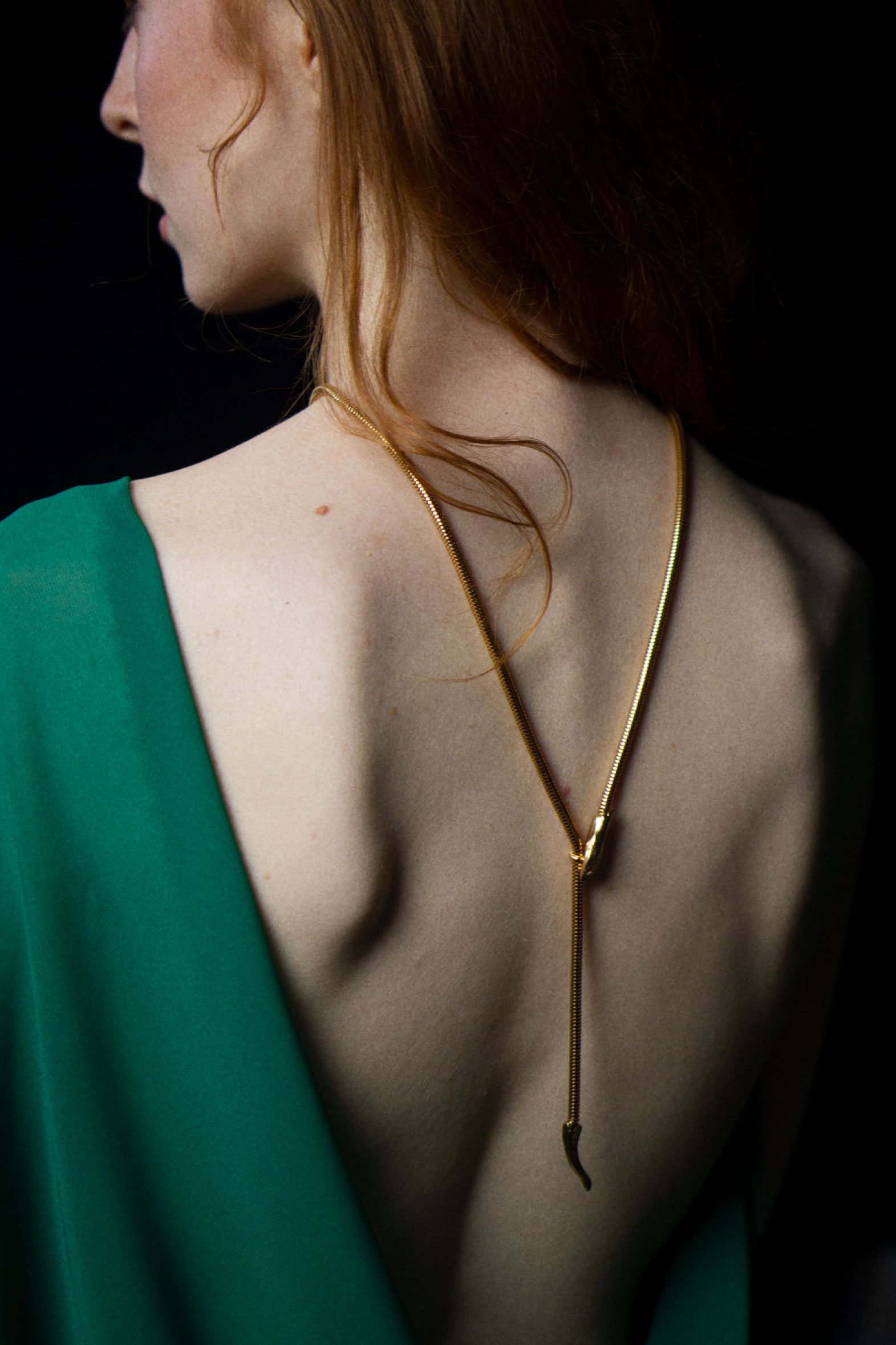 Elegant portrait of a red-haired woman's back, showcasing a gold necklace and a draped green fabric against a dark backdrop.