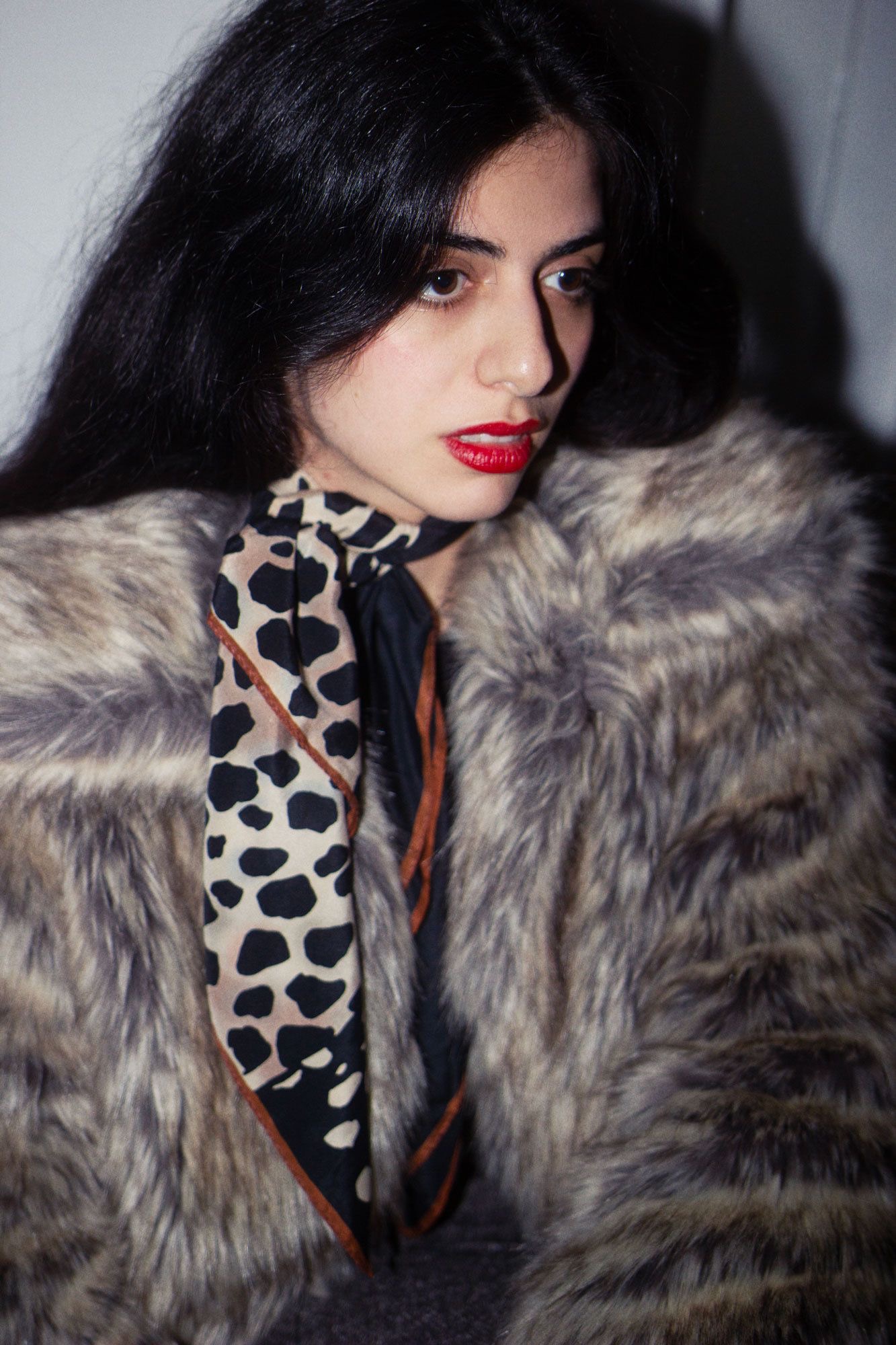 Portrait of a young woman with dark hair, wearing a fur coat and animal print scarf, looking off camera. Red lipstick.