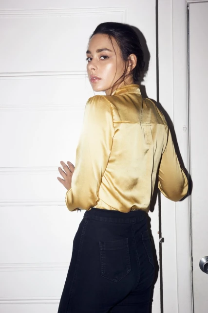 A young woman with dark hair, wearing a gold blouse and black pants, stands indoors near a white door, looking over her shoulder at the camera.