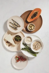 Top-down view of ceramic plates arranged on a light background, featuring sardines, papaya, cheese, asparagus, beans, and olives.
