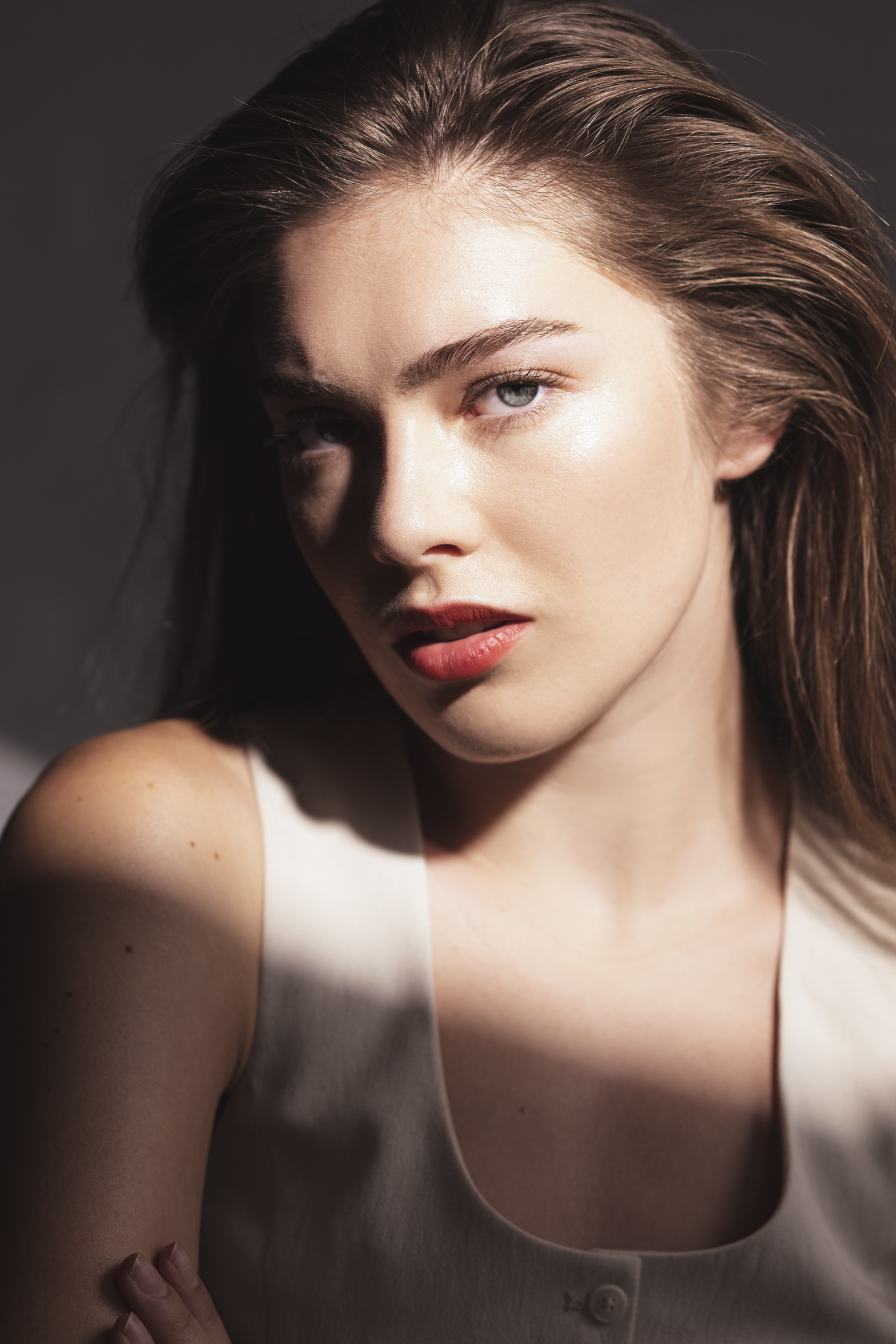 Close-up portrait of a young adult female with brown hair and a light complexion, wearing a sleeveless top against a grey background.