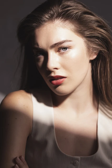 Close-up portrait of a young adult female with brown hair and a light complexion, wearing a sleeveless top against a grey background.