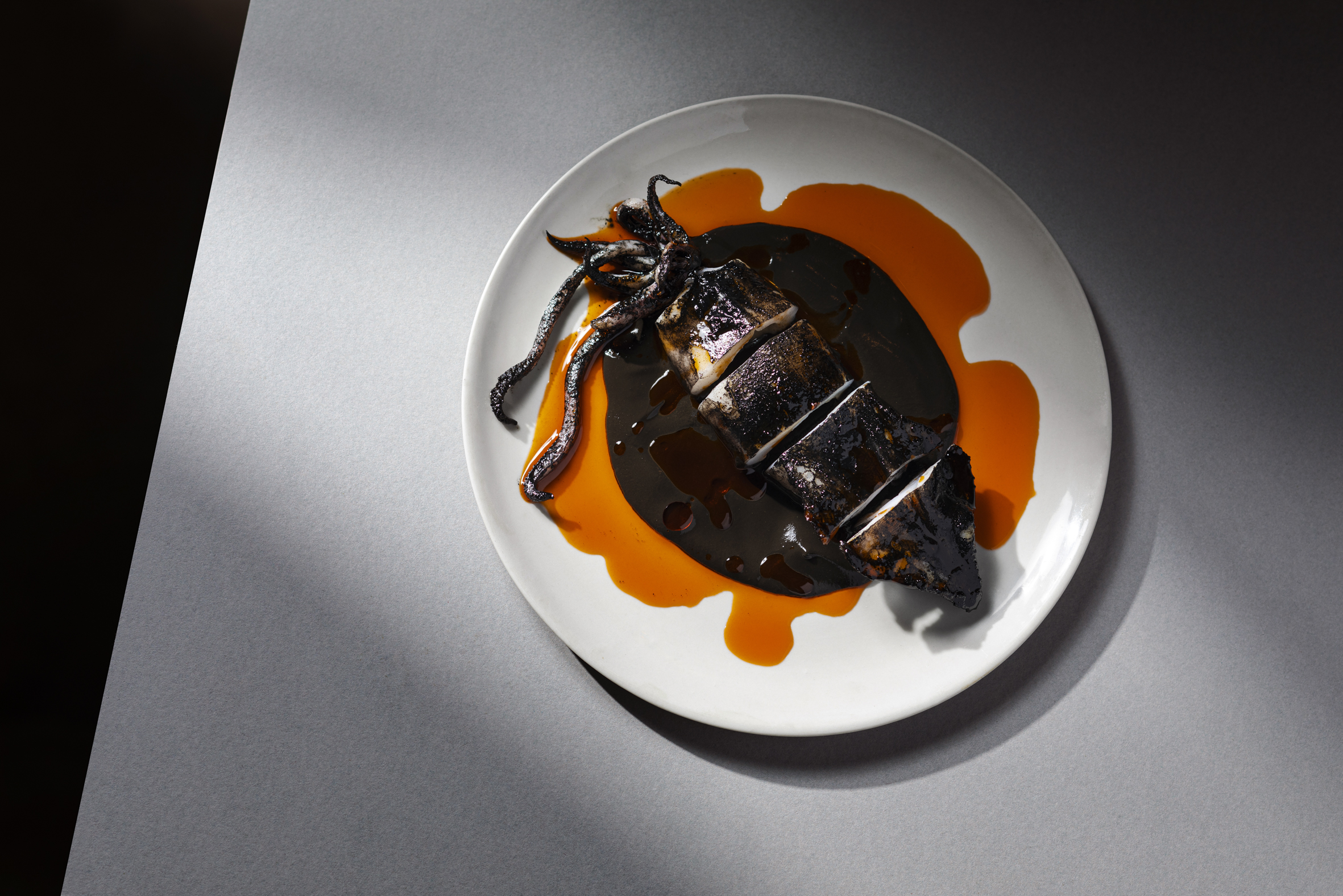 Overhead shot of grilled squid on a white plate, drizzled with black and orange sauce on a gray surface with lighting accents.