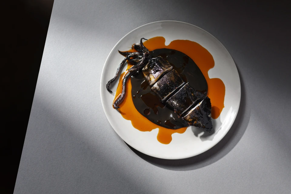 Overhead shot of grilled squid on a white plate, drizzled with black and orange sauce on a gray surface with lighting accents.