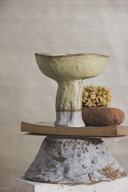 Still life of a pale yellow ceramic pedestal bowl on top of a stack of book, orange rock, and light grey rock against a beige background.