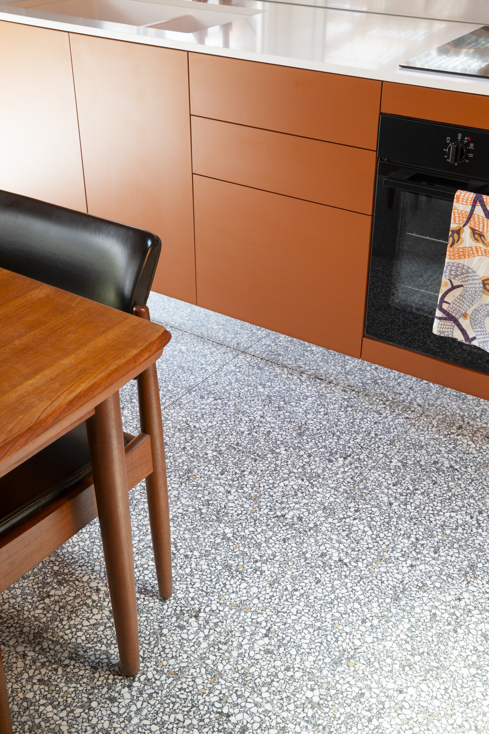 Interior shot showcases modern kitchen with orange cabinetry, light terrazzo floor, and a wooden table with black vinyl chair.
