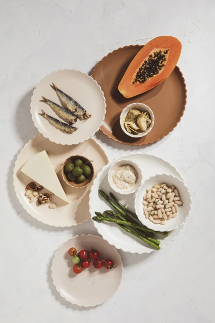 Overhead shot of colorful food plates featuring sardines, papaya, tomatoes, asparagus, cheese, beans, olives, and sauce on a white surface.