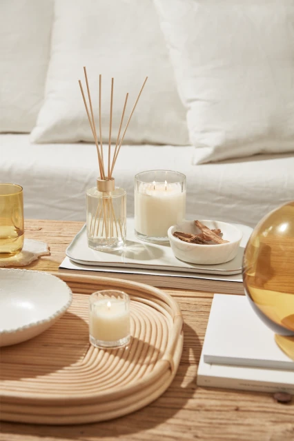 Still life featuring a reed diffuser, candles, palo santo in a small bowl, and decorative glass orb on a wooden table with white linens.