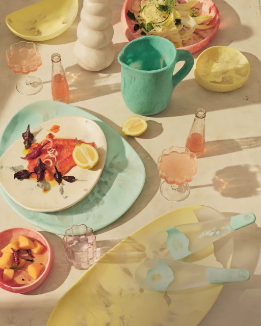 Still life with salmon dish, sliced lemon, pink drink, and assorted pastel ceramic dishes and serving-ware arranged on a table.
