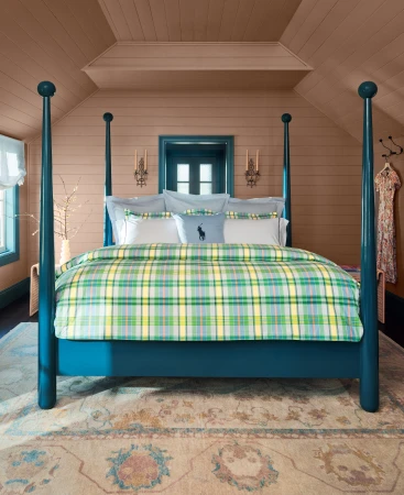 Bedroom features a blue four-poster bed with plaid blanket, patterned rug, tan-colored walls, white pillows, and sconces. 