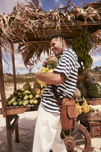 Smiling man holding fresh fruit at an outdoor market stand on a sunny day, wearing a striped shirt and light pants, with a beach background.