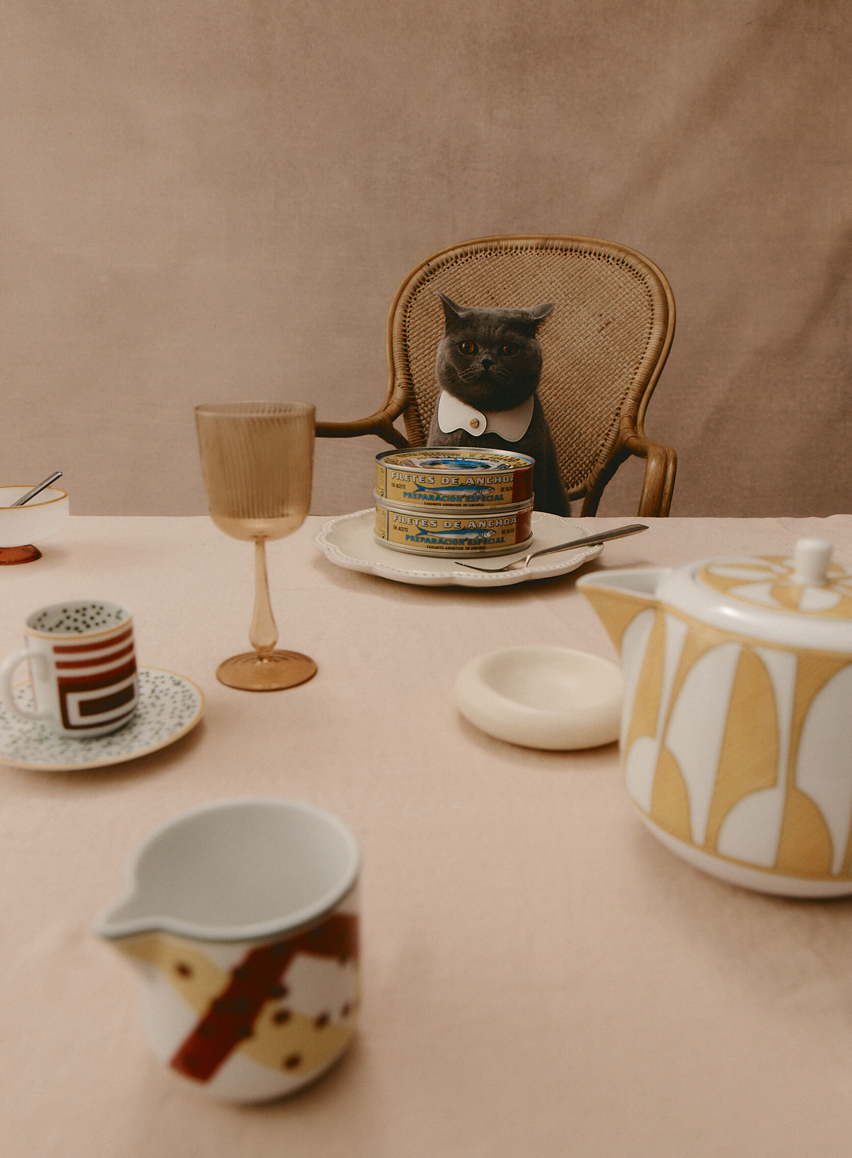 A gray cat wearing a white bow tie sits at a dining table with plates, glasses, and canned goods, creating a humorous still life.