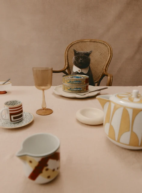 A gray cat wearing a white bow tie sits at a dining table with plates, glasses, and canned goods, creating a humorous still life.