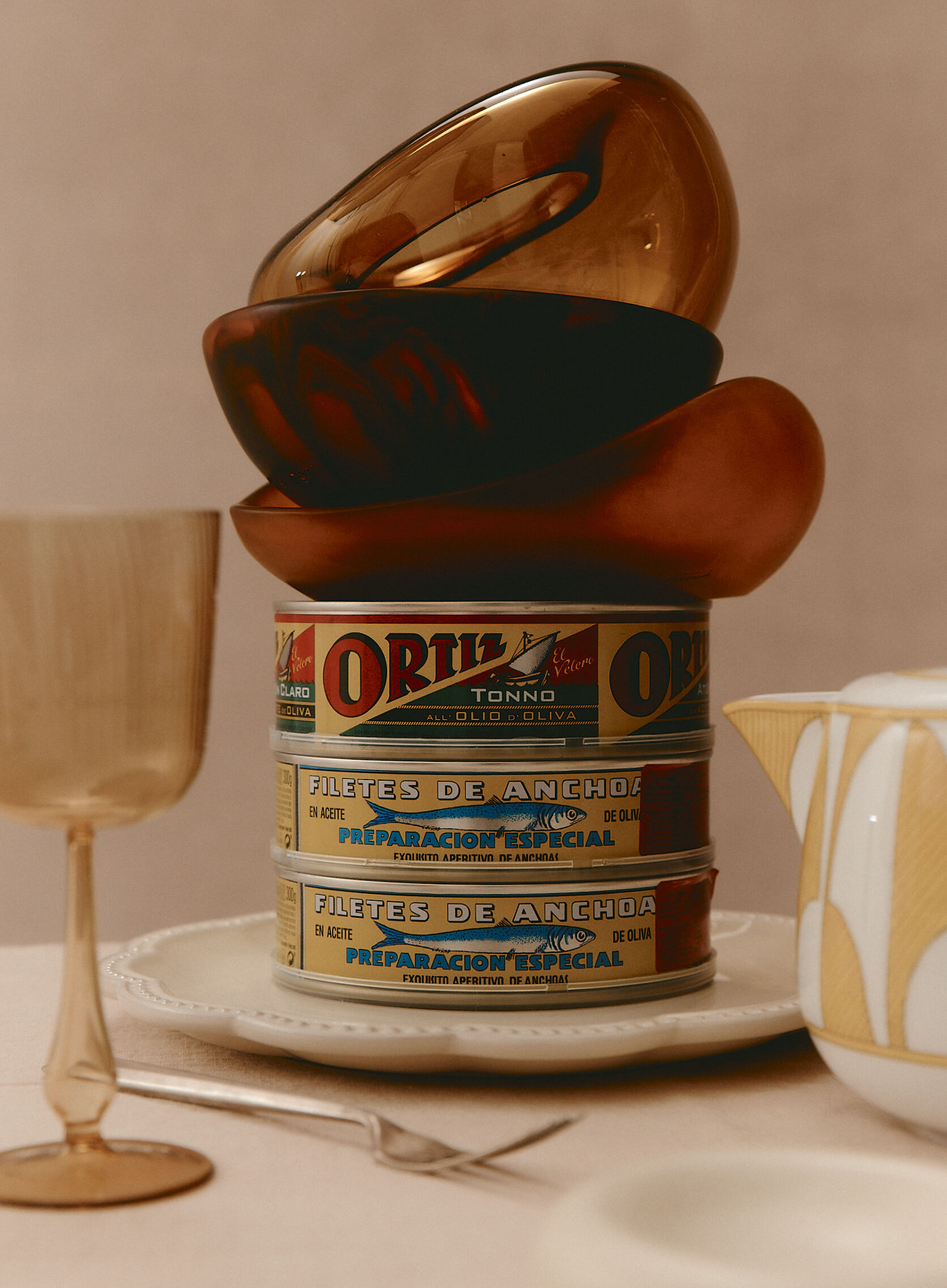 Still life composition featuring a stack of three Ortiz tuna cans, topped with wooden bowls, arranged on a white plate near a golden glass and teapot.