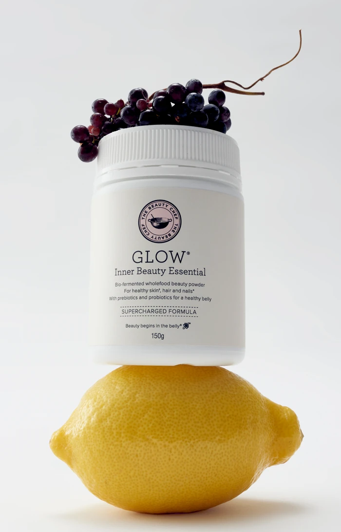 White supplement jar balanced on a yellow lemon, topped with dark grapes against a plain white background.