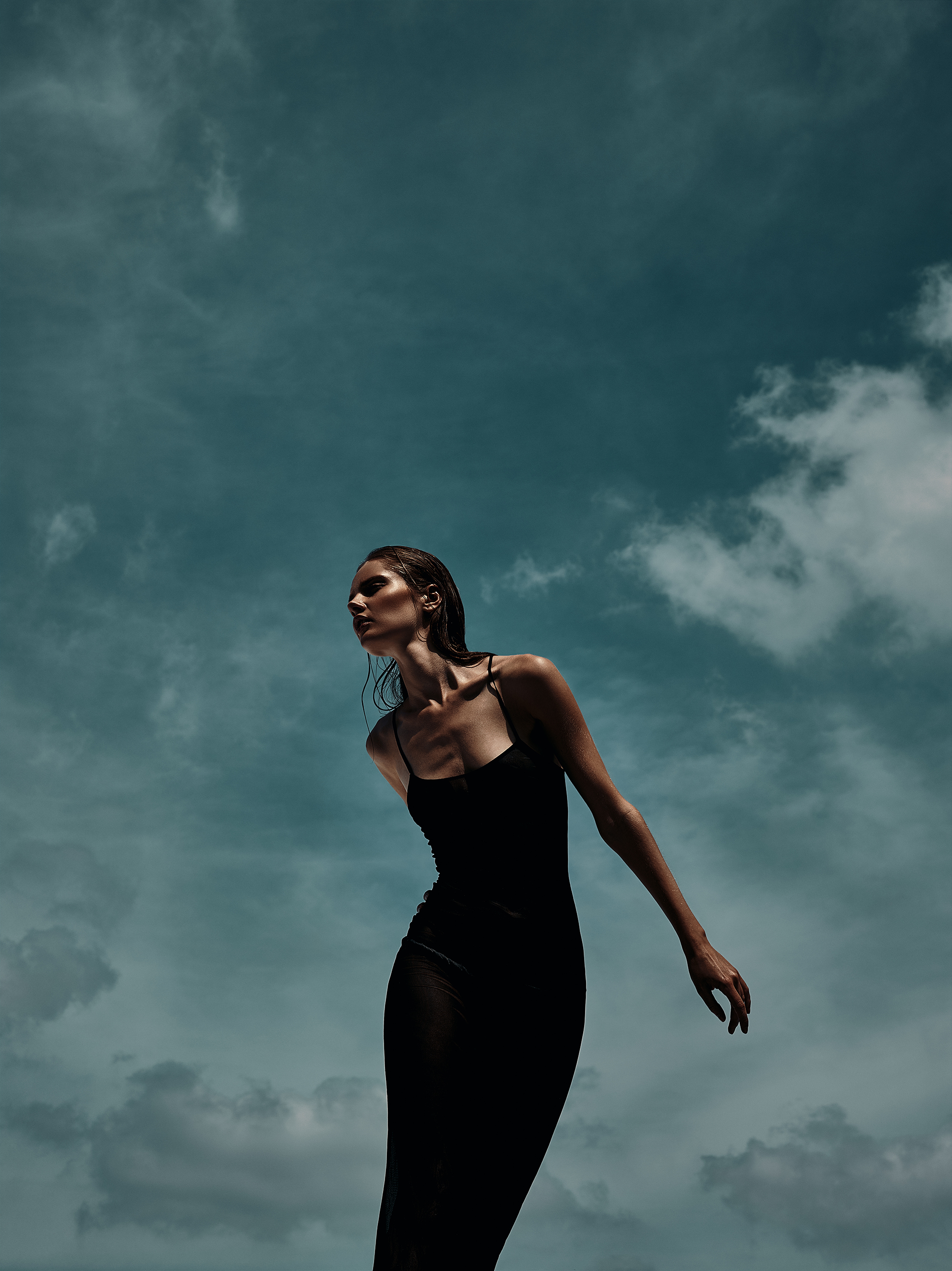 A slender woman in a black dress stands against a cloudy sky, illuminated by bright light, looking off-camera in a fashion editorial shot.