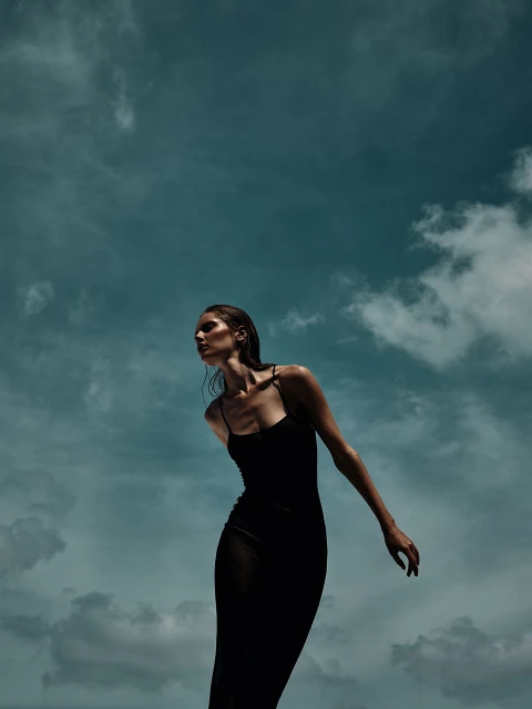 A slender woman in a black dress stands against a cloudy sky, illuminated by bright light, looking off-camera in a fashion editorial shot.