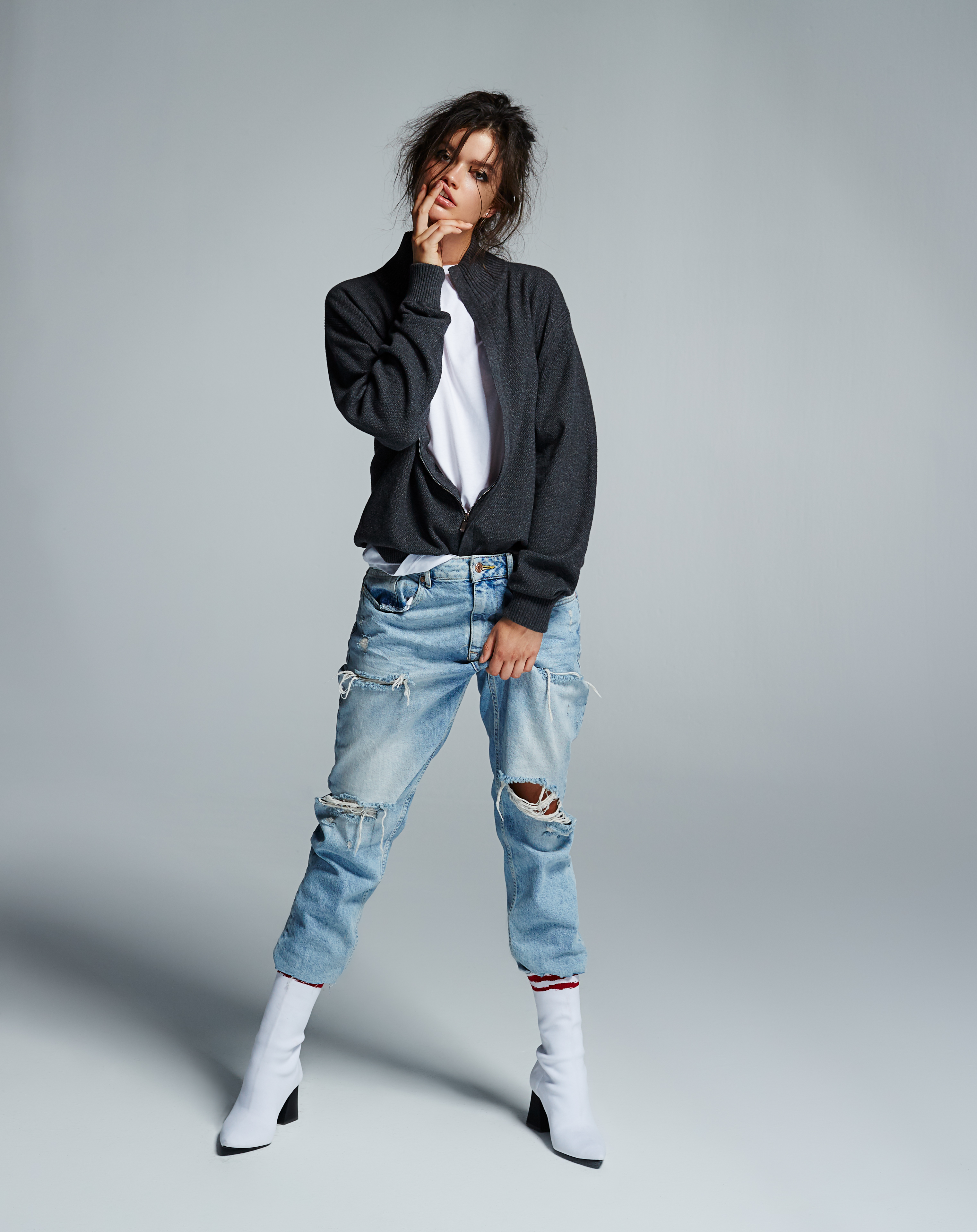 Woman in ripped jeans, gray jacket, and white boots poses in a studio with neutral lighting. Fashion editorial portrait.
