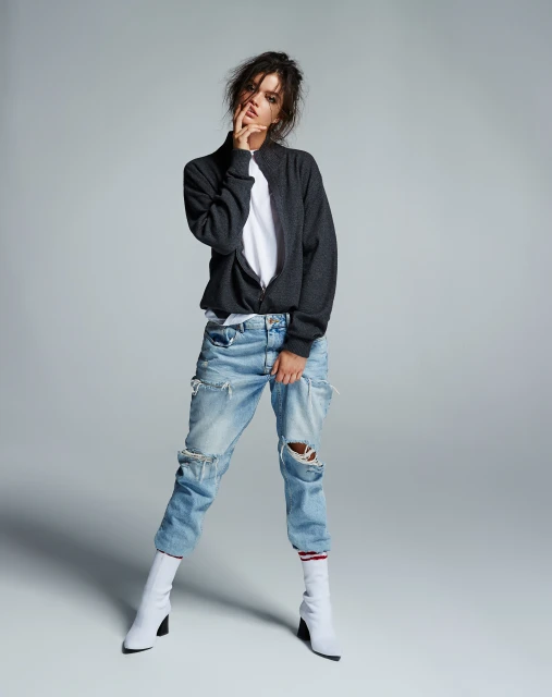 Woman in ripped jeans, gray jacket, and white boots poses in a studio with neutral lighting. Fashion editorial portrait.