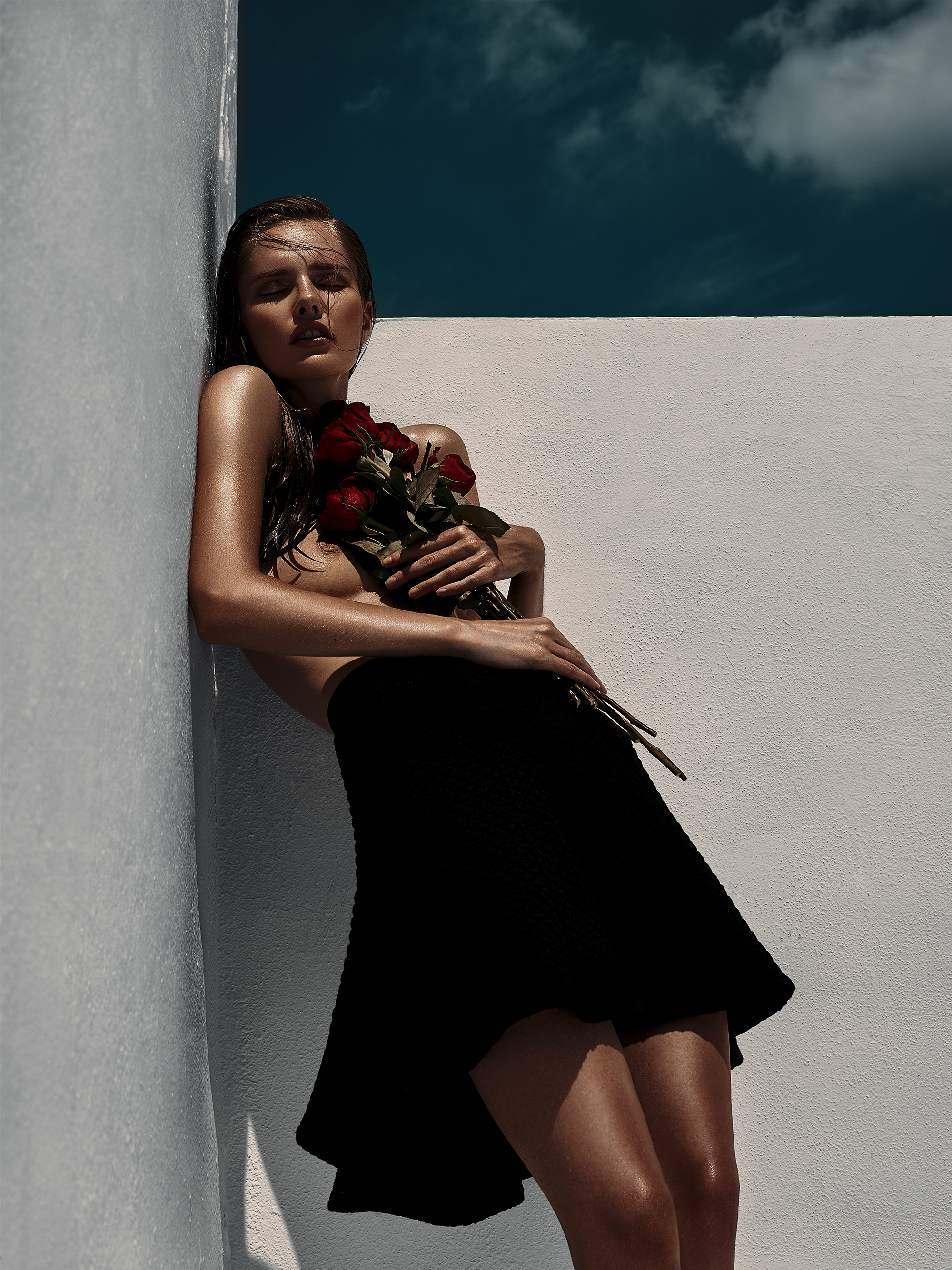 A fair-skinned woman with wet hair, wearing a black skirt and holding red roses, leans against a white wall under a partly cloudy blue sky.
