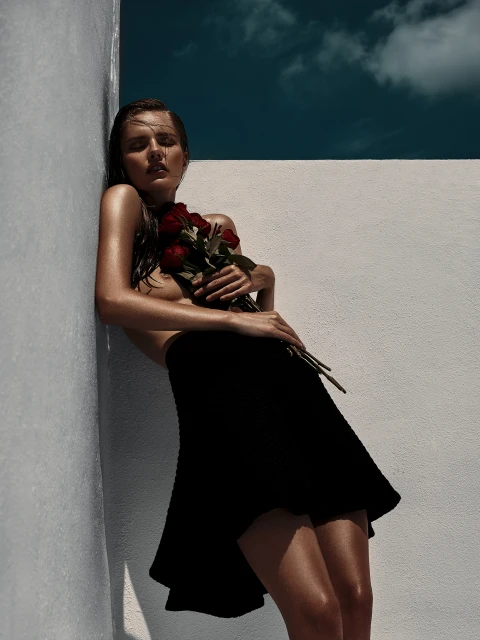 A fair-skinned woman with wet hair, wearing a black skirt and holding red roses, leans against a white wall under a partly cloudy blue sky.