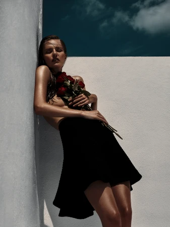 A fair-skinned woman with wet hair, wearing a black skirt and holding red roses, leans against a white wall under a partly cloudy blue sky.
