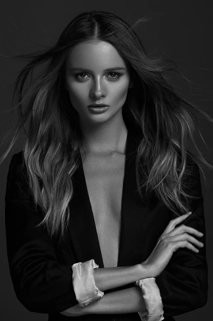 Monochrome studio portrait of a stylish adult woman with long hair in a blazer with arms crossed and a neutral facial expression.