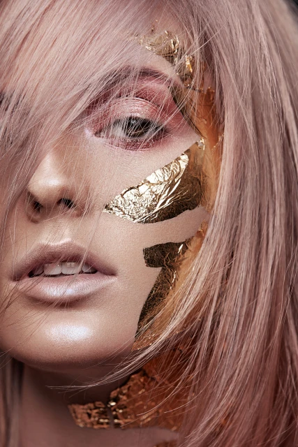 Close-up portrait of a stylish young adult woman with pink hair and gold leaf on her face, gazing directly at the camera, in a beauty shot.