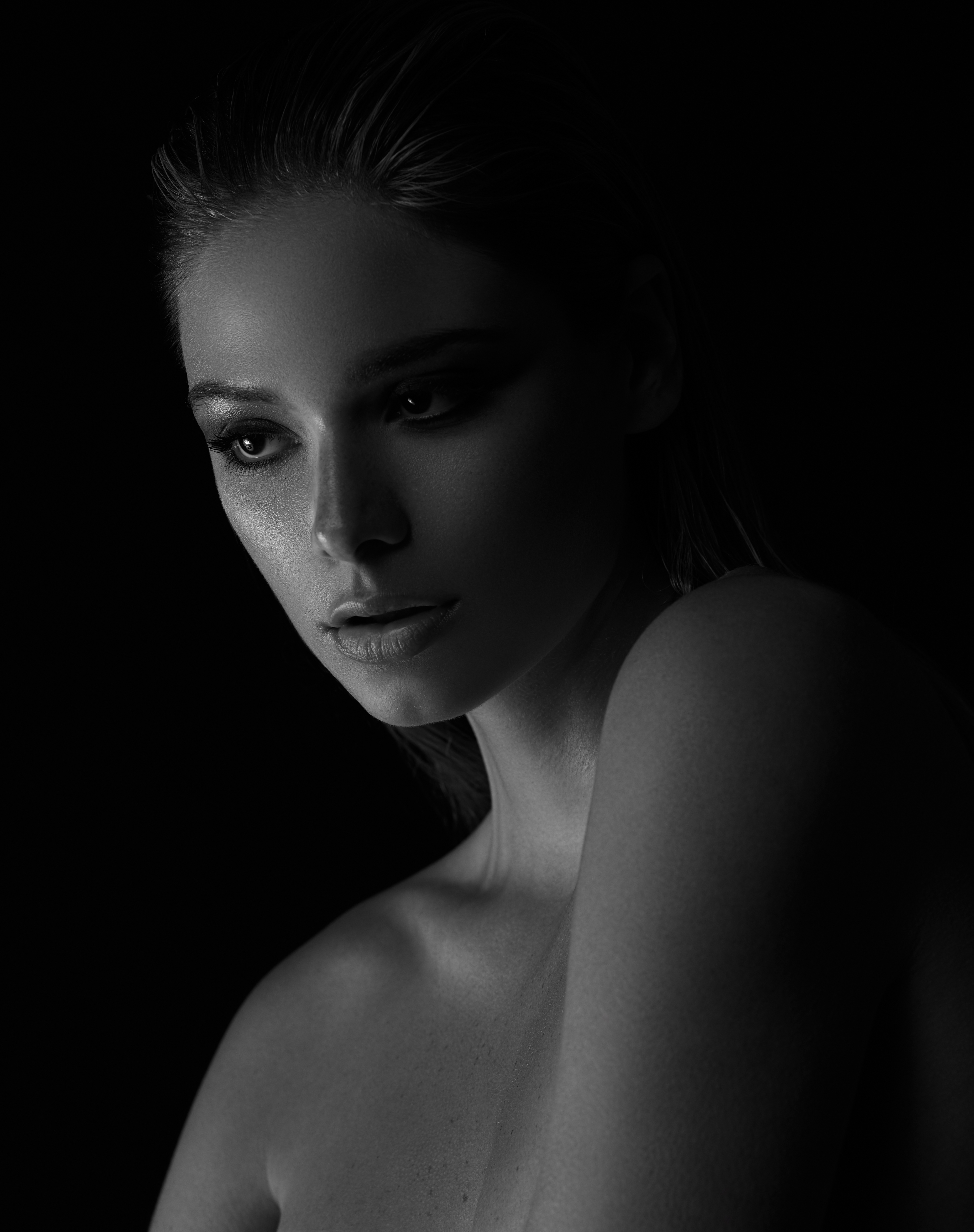 Monochrome portrait of a fair-skinned adult woman with wet hair and dark eye makeup against a black backdrop, looking downward.