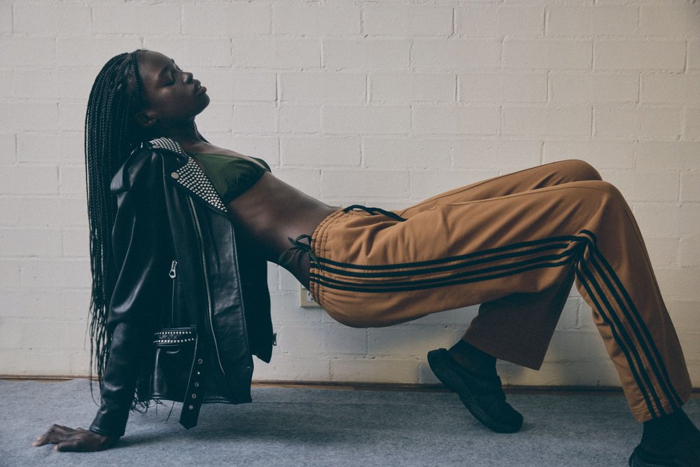 Woman in stylish sportswear and studded leather jacket poses against white brick wall, eyes closed, fashion studio, medium shot.