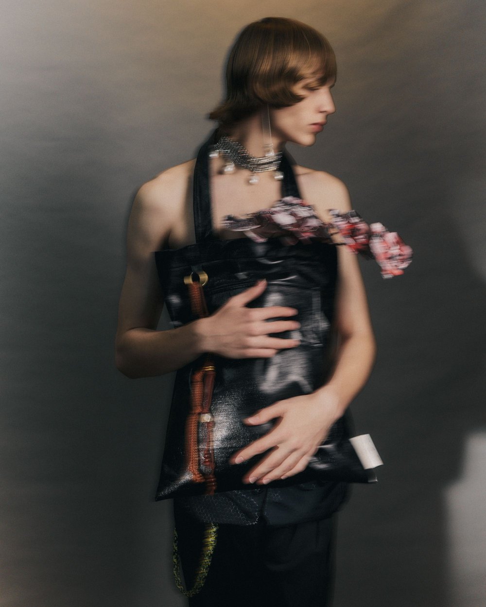 Person with short brown hair, adorned with a silver necklace and earrings, poses with a black bag holding flowers.